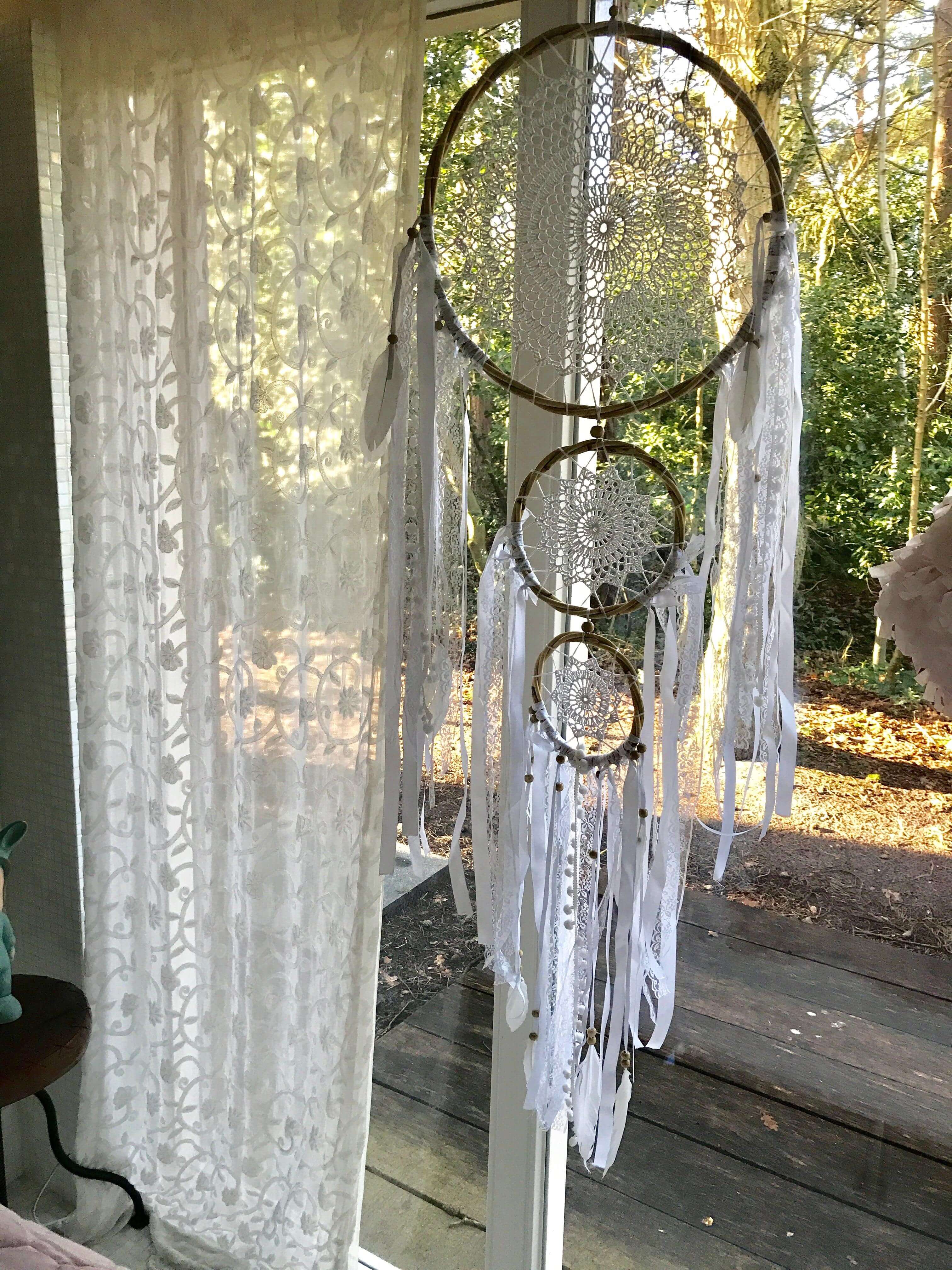 Large white dreamcatcher trio with ribbons, laces, and pompoms hanging indoors near window.