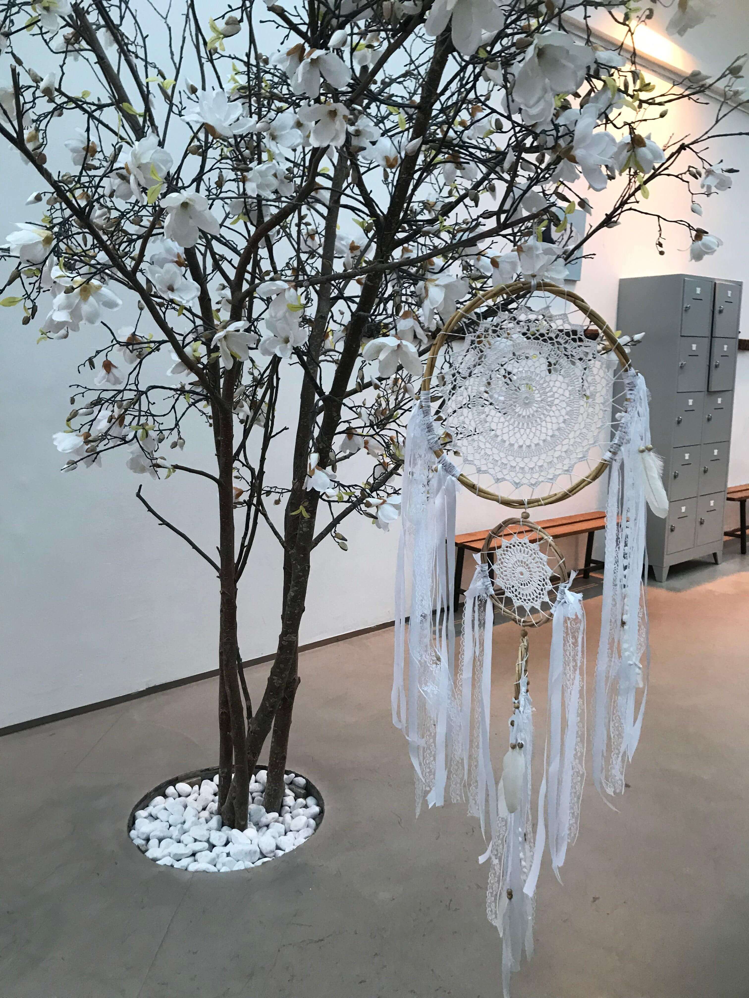 White handmade dream catcher with intricate lace center, long white ribbons, and feathers hanging from a branch with small.