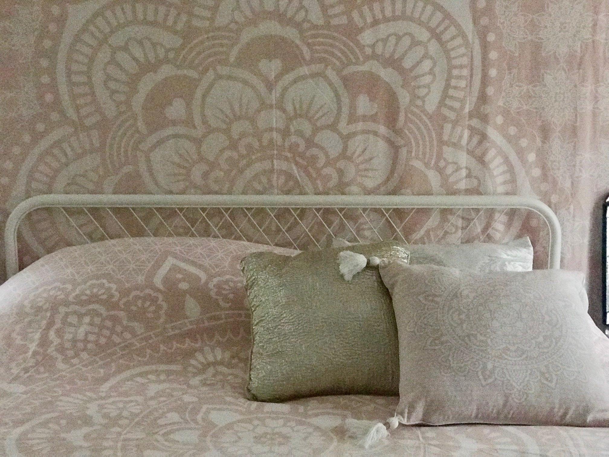 Pink bed cover with intricate mandala pattern draped over white metal bed frame, two textured throw pillows in front.