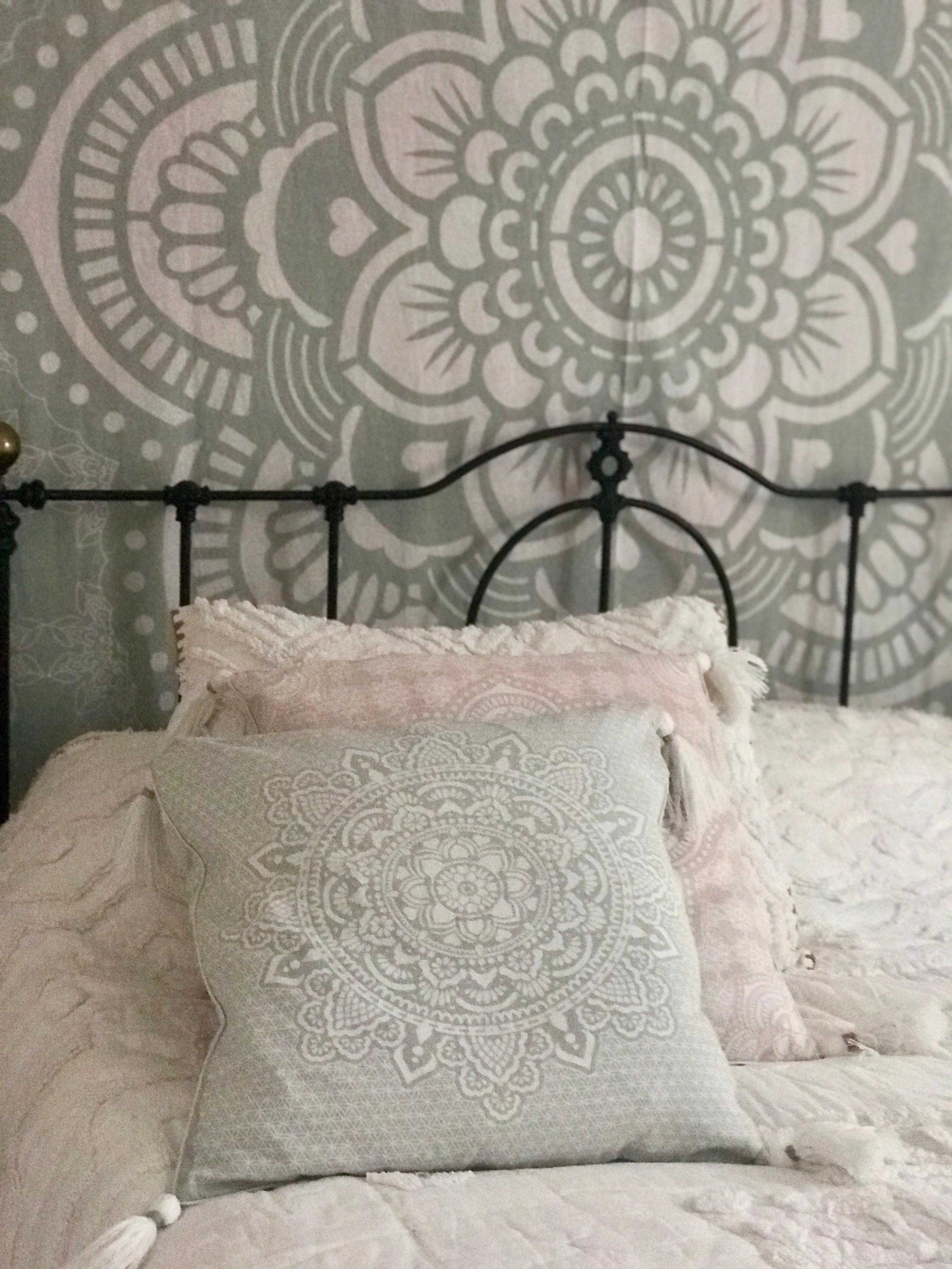 Mint green square cushion with intricate holy mandala embroidery on bed with soft pink pillows and black metal headboard.