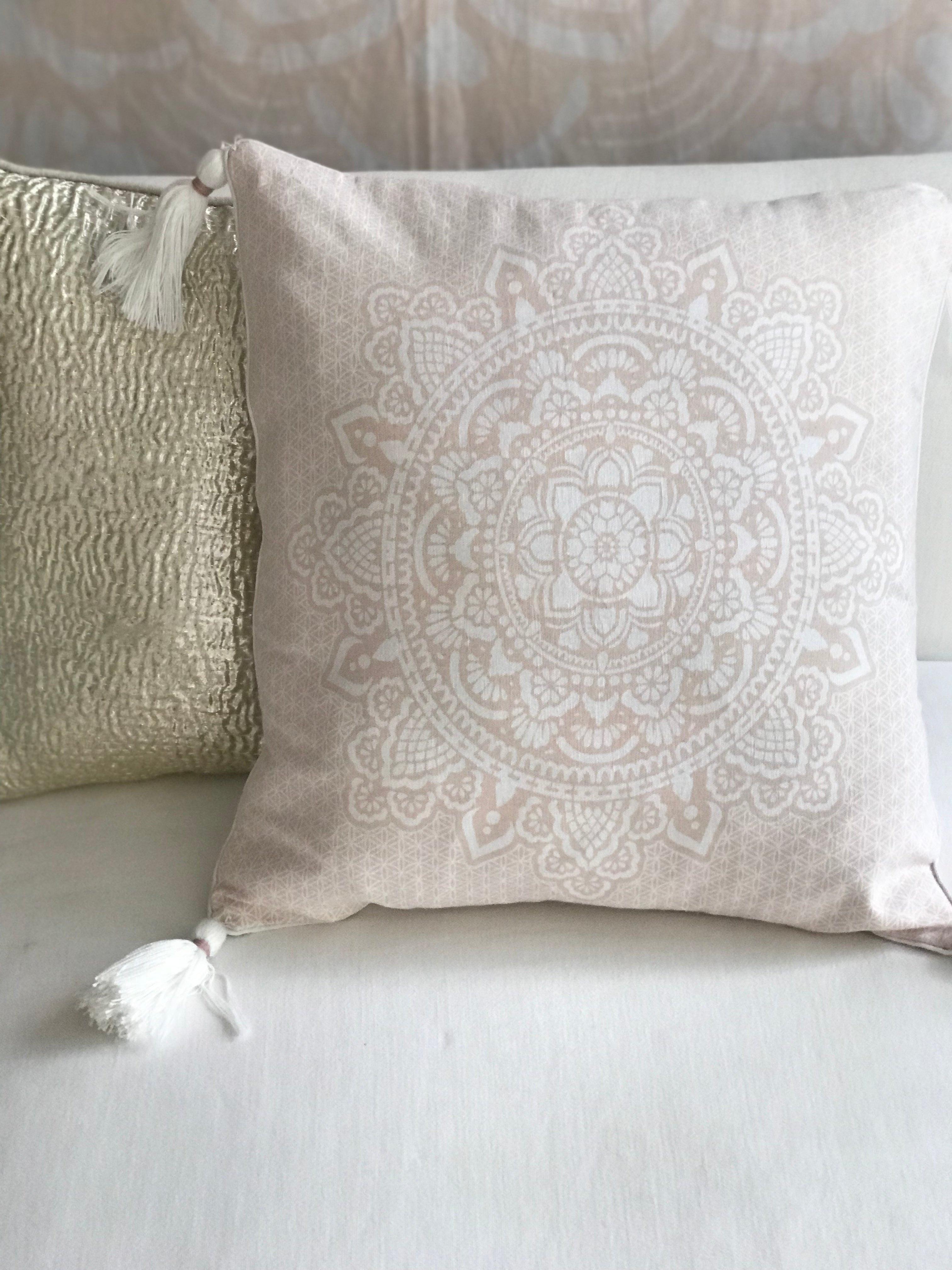 Square pink cushion with white intricate mandala design and tassels on each corner.