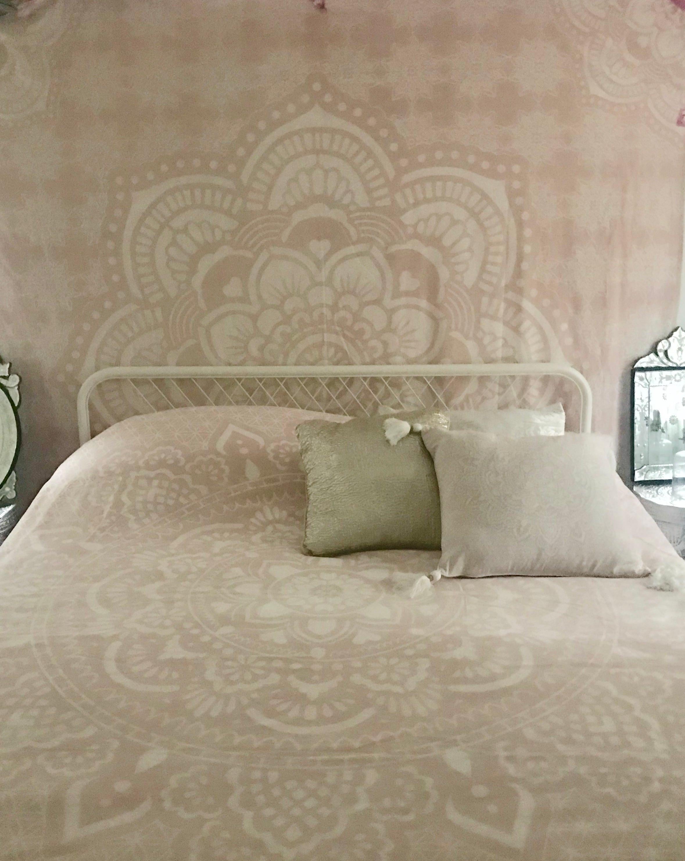 Soft pink bed cover with intricate Holy Mandala design and two neutral throw pillows on a white metal bed frame.