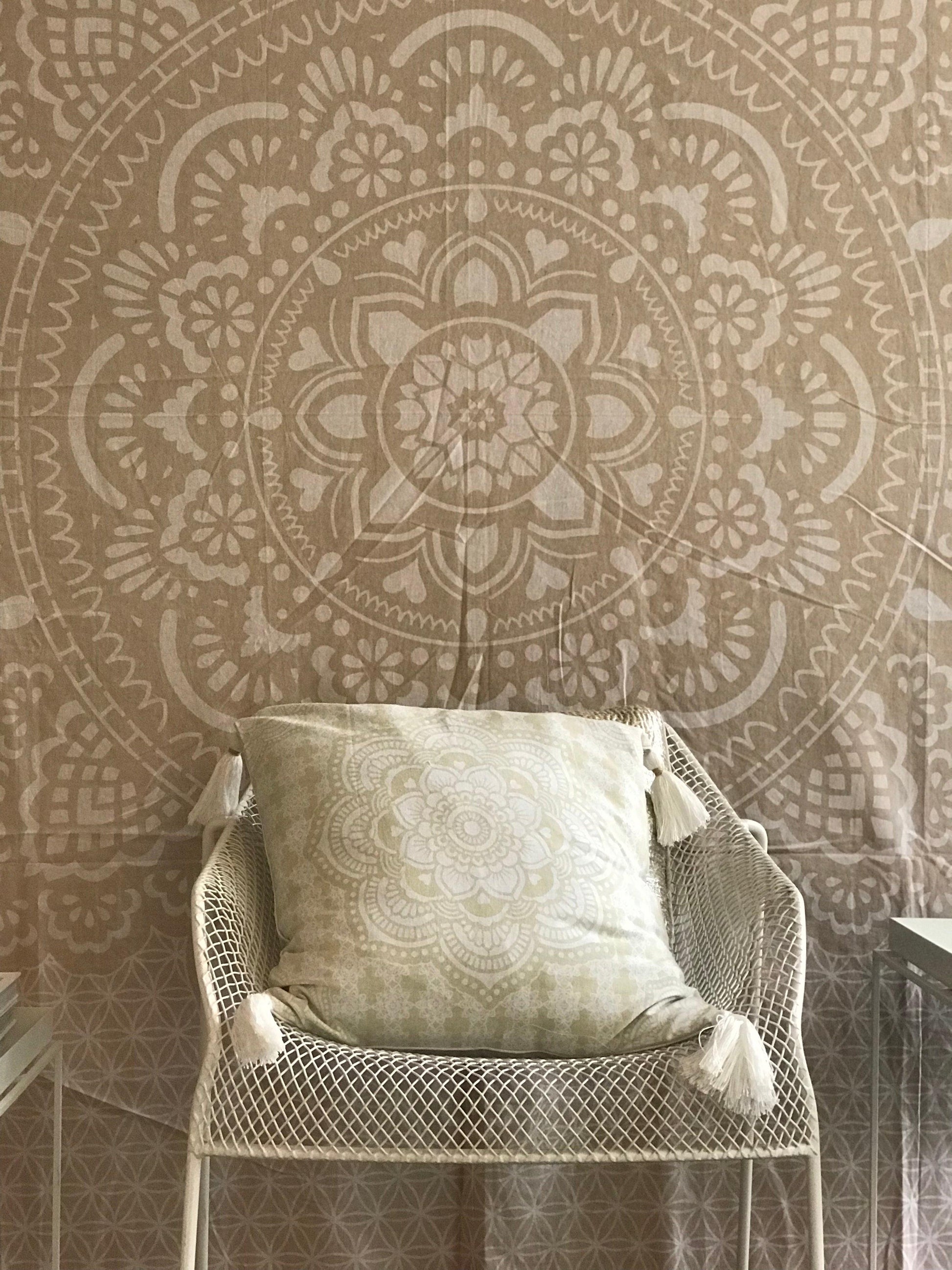 Khaki tapestry with intricate holy mandala design in beige tones behind woven chair with light beige cushion.