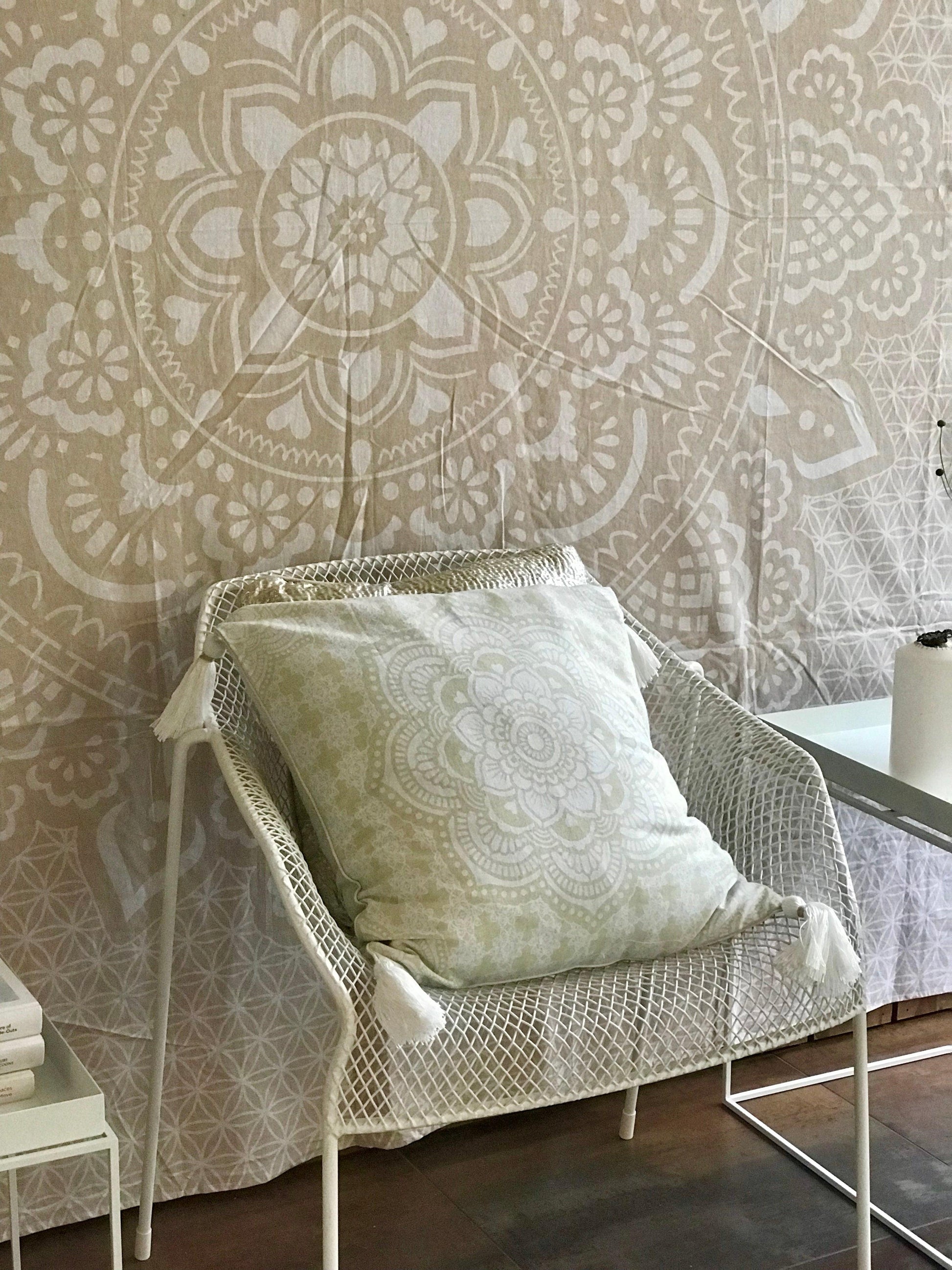 Khaki tapestry with intricate holy mandala pattern hanging behind a white wicker chair with a matching cushion.