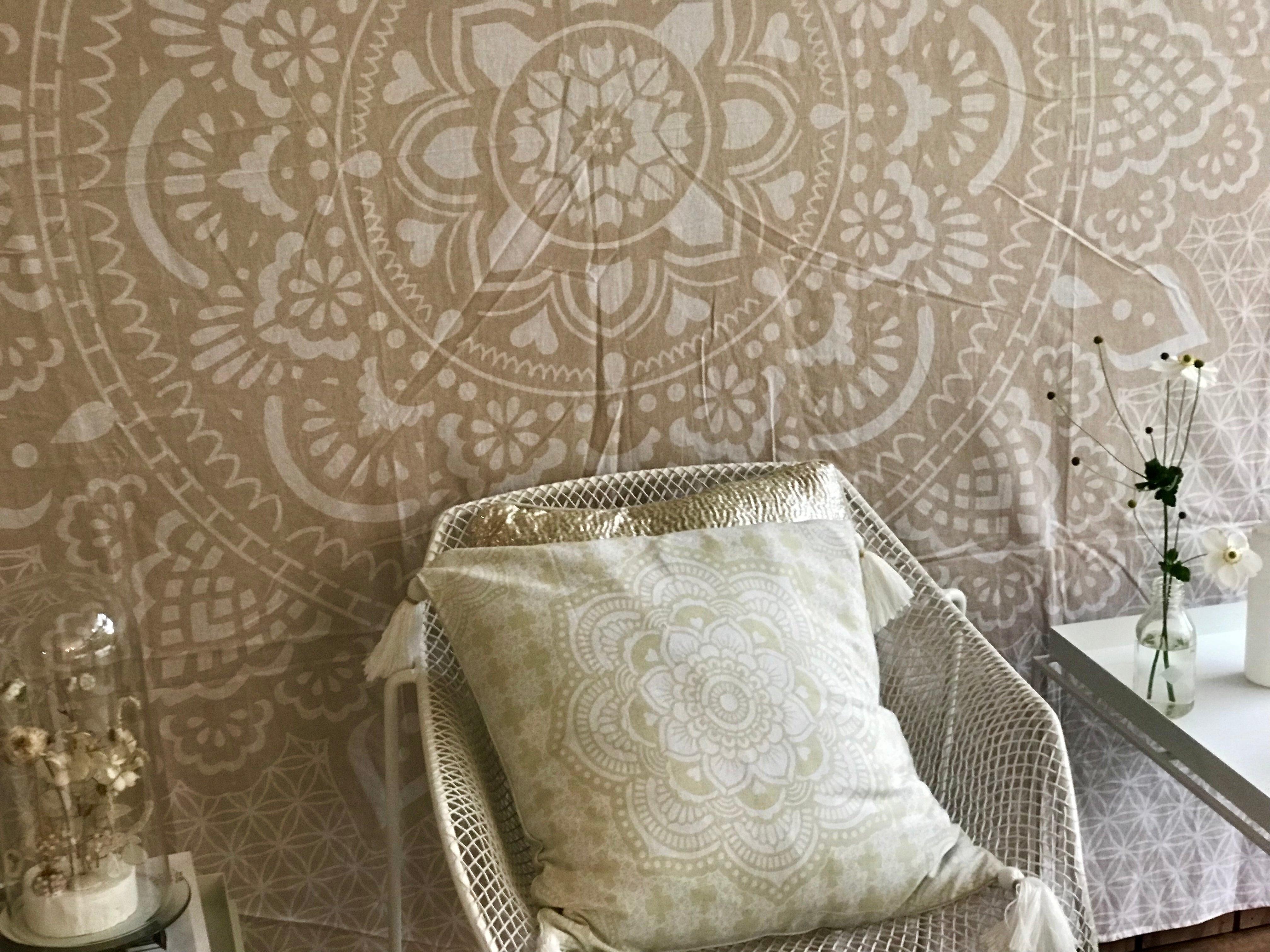 Khaki tapestry with intricate holy mandala design hanging on wall behind white wicker chair and matching cushion.