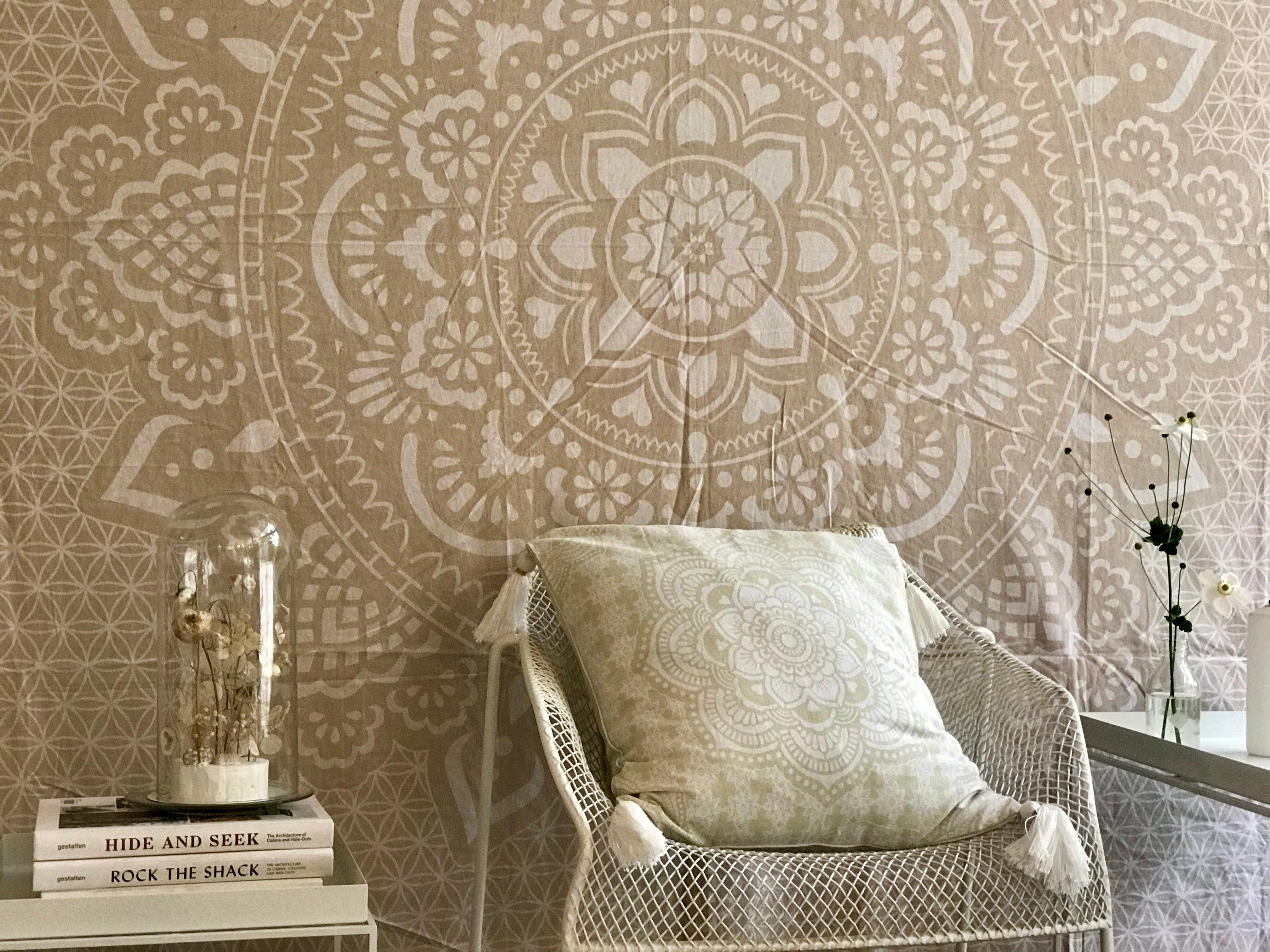 Khaki tapestry with intricate holy mandala pattern displayed on a wall behind a white wire chair with a beige patterned.