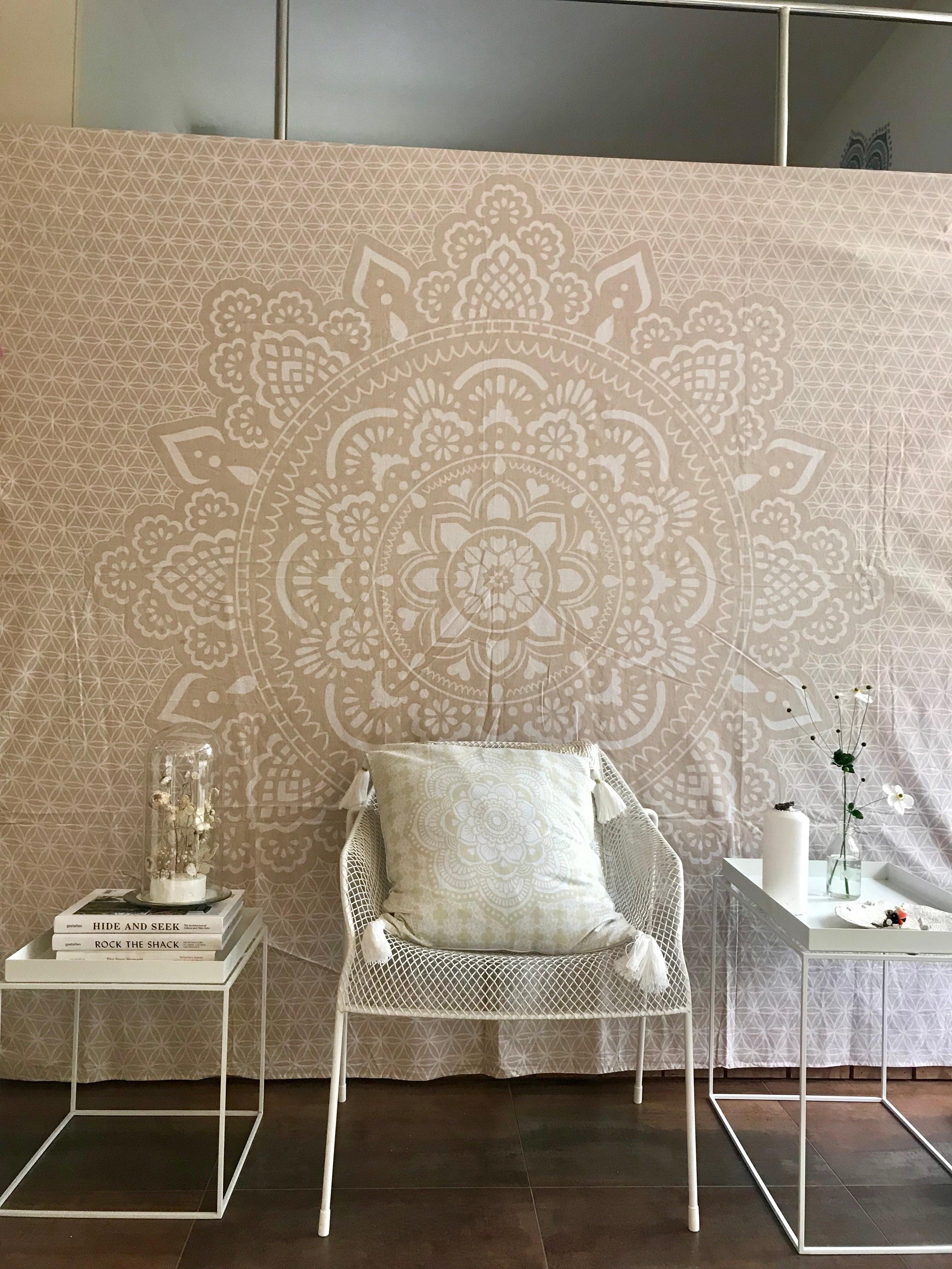 Khaki tapestry with intricate white mandala design hanging behind white metal chair and white side tables.