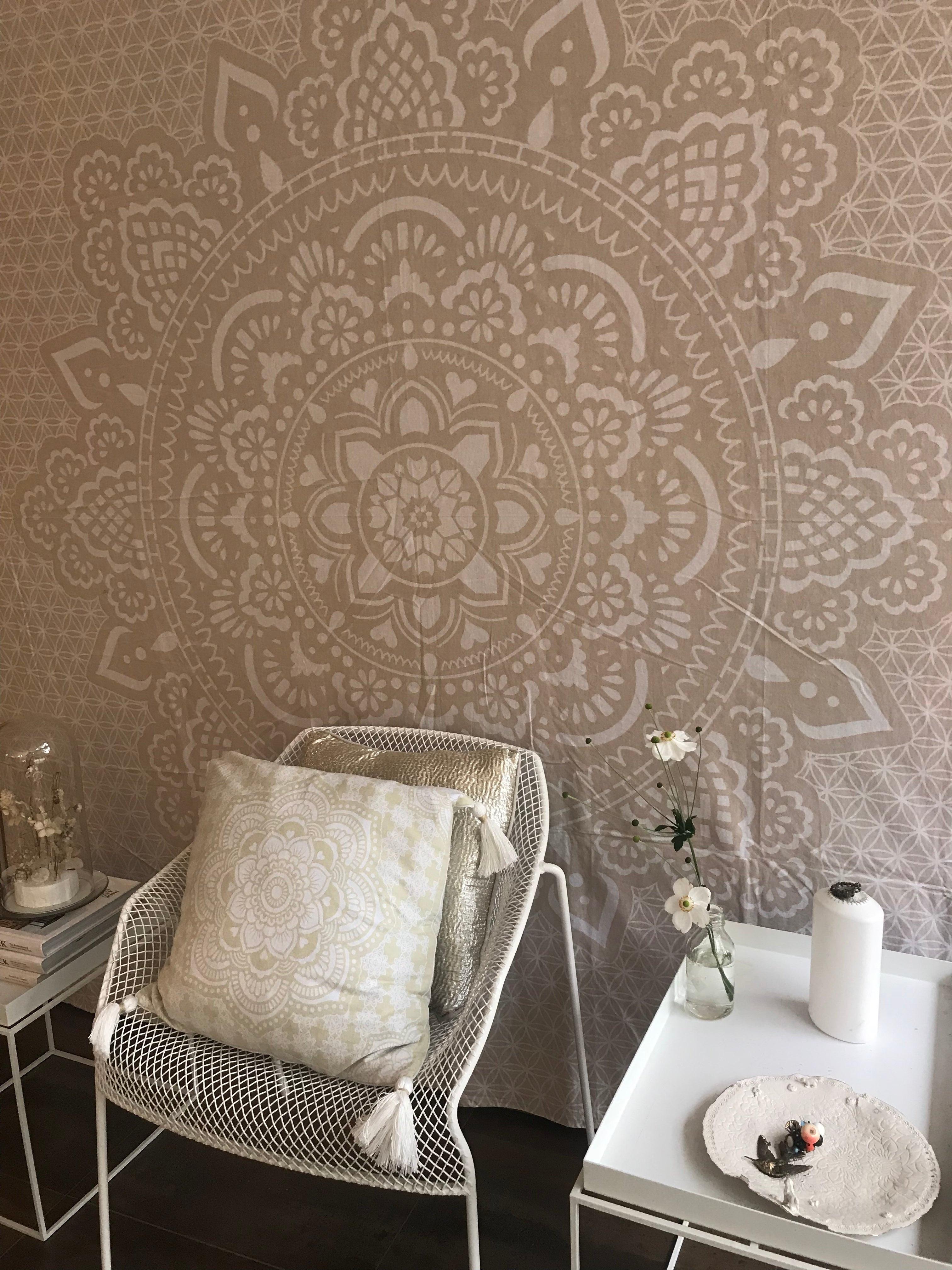 Khaki tapestry with intricate white mandala design hanging above white metal chair with beige cushion in modern room setting.