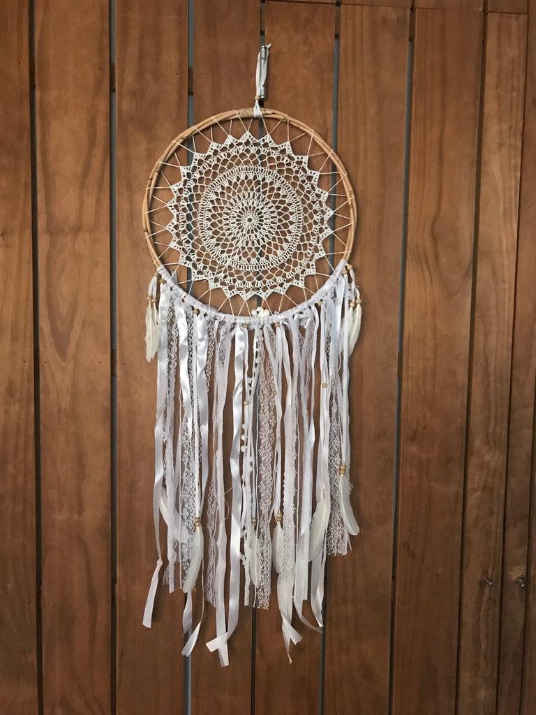 White crochet dreamcatcher 42cm with intricate circular web and long flowing ribbons against wooden wall.