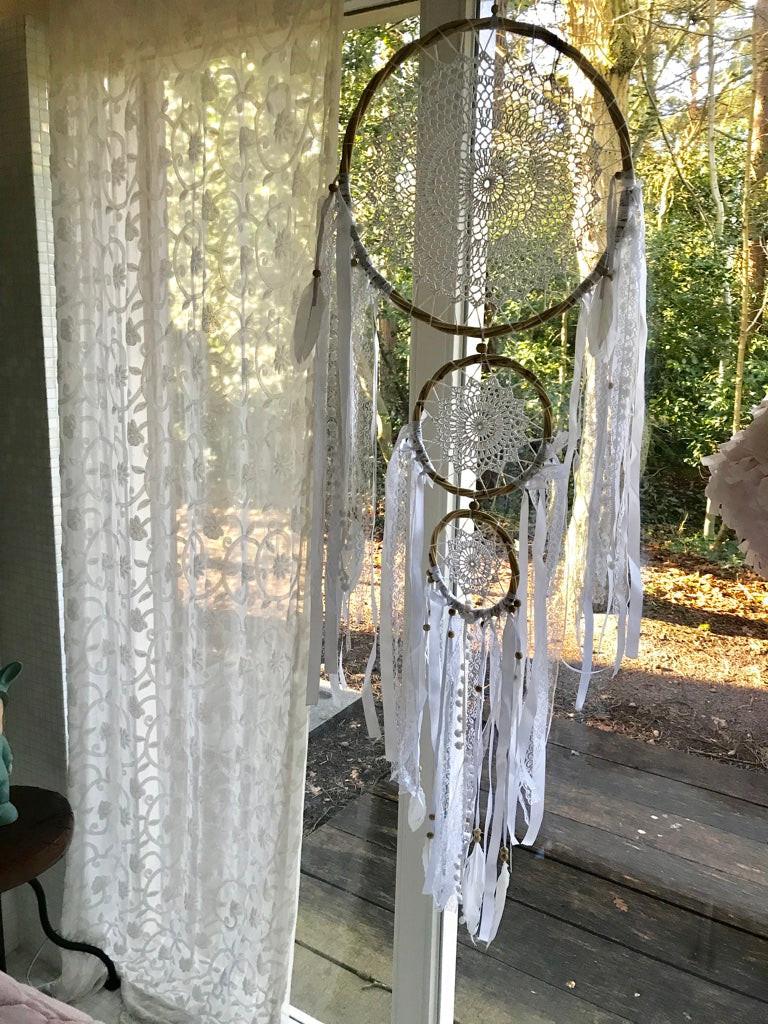 Three-tier dreamcatcher with brown hoops and white feathers hanging by a window with lace curtains and outdoor view.