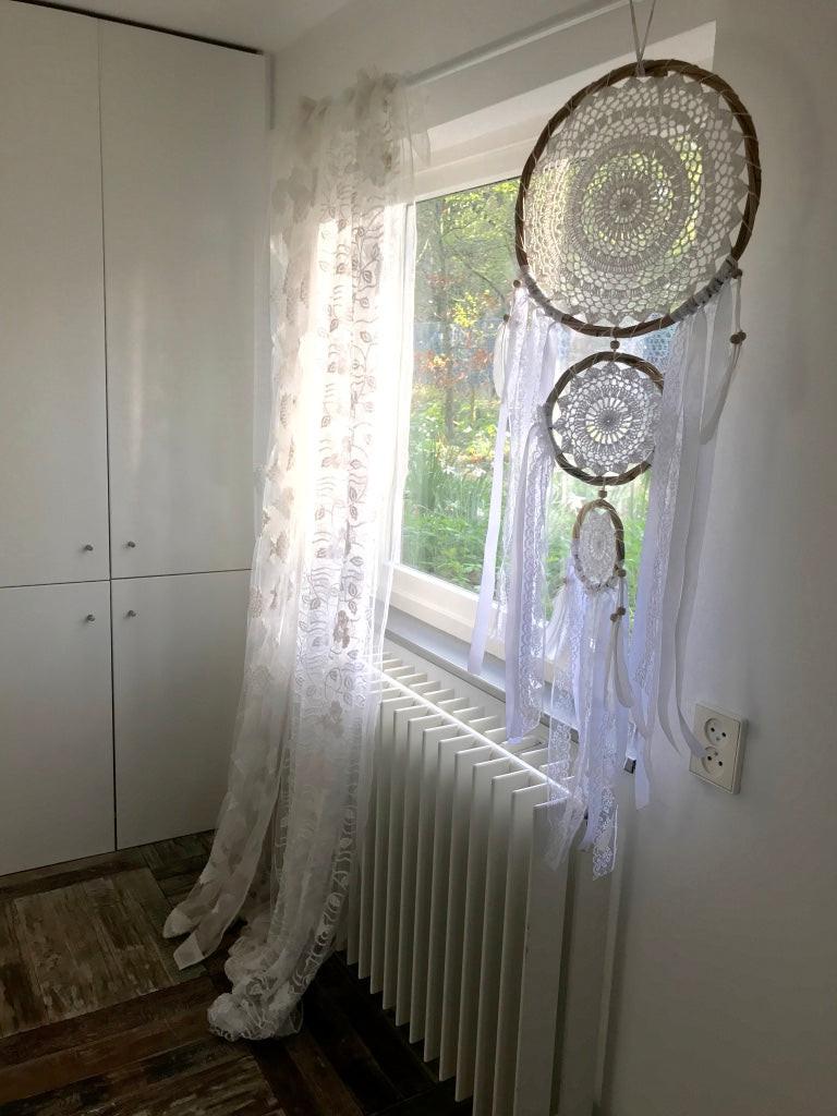 Boho dream catcher trio with white feathers and lace hanging by sunlit window.